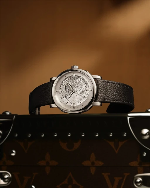 Elevated view of luxury wristwatch with black leather band on branded Louis Vuitton trunk against brown backdrop in studio lighting.