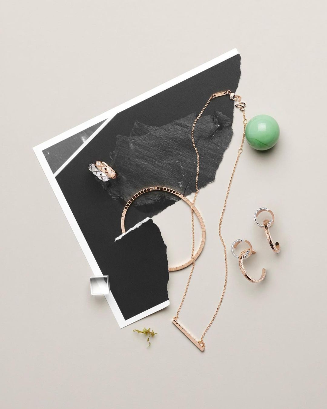 Rose gold jewellery pieces arranged on a neutral surface with torn paper, textured black fragments, and a green sphere