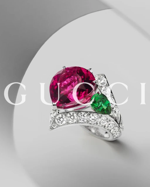 Close-up of a gemstone ring with a large pink stone, accented by white diamonds and a green stone, set against a soft grey backdrop.