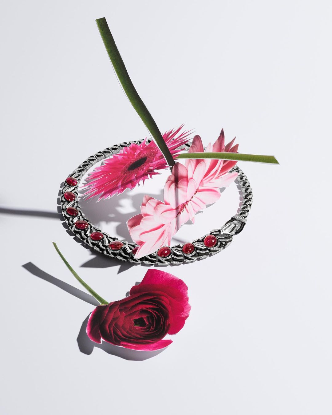 Silver bracelet with red gemstones surrounded by pink gerbera daisies and a red ranunculus flower on a white surface, studio lighting.