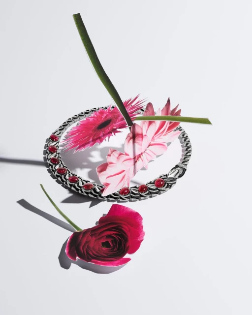 Silver bracelet with red gemstones surrounded by pink gerbera daisies and a red ranunculus flower on a white surface, studio lighting.