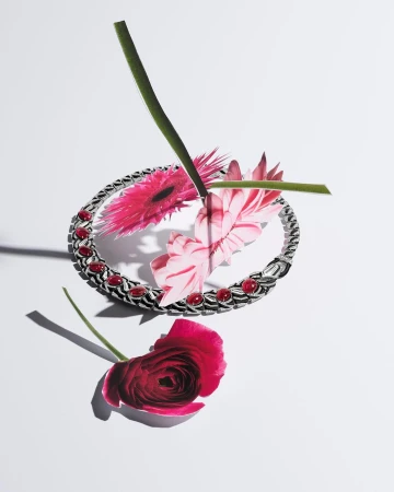 Silver bracelet with red gemstones surrounded by pink gerbera daisies and a red ranunculus flower on a white surface, studio lighting.