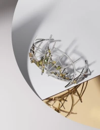 Detailed silver lattice tiara embellished with green and yellow gemstones resting against a curved white and gold background.
