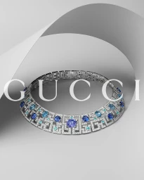 An ornate silver gemstone necklace set with blue sapphires on a curved white seamless studio backdrop with Gucci text overlay.
