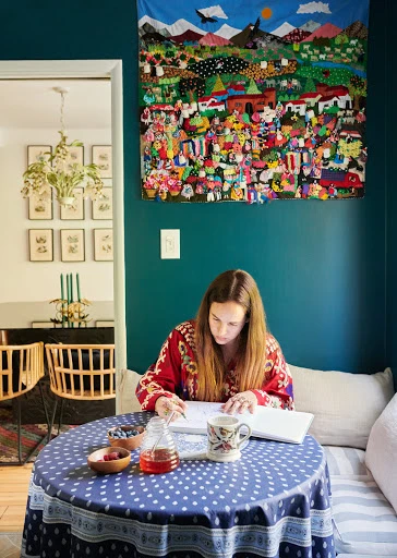 Woman sits at a table, writing in a notebook, with a cup of tea, fruit, and colorful textile art on the wall in an interior setting.