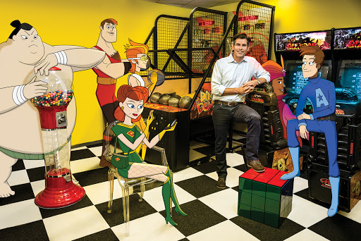 Animated characters and a man pose in an arcade with a gumball machine, including a sumo wrestler, super hero, and video games.