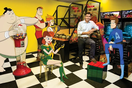 Animated characters and a man pose in an arcade with a gumball machine, including a sumo wrestler, super hero, and video games.
