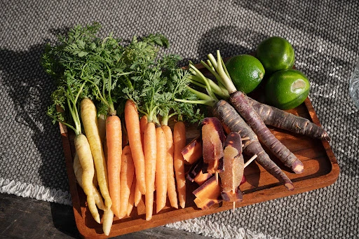 Still life of colorful carrots (yellow, orange, purple) with fresh green tops, limes, and carrot peels on a wooden tray.