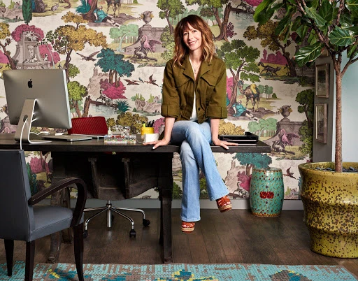 Woman sitting on desk in home office with desktop computer, colorful scenic wallpaper, potted tree, and hardwood floor.