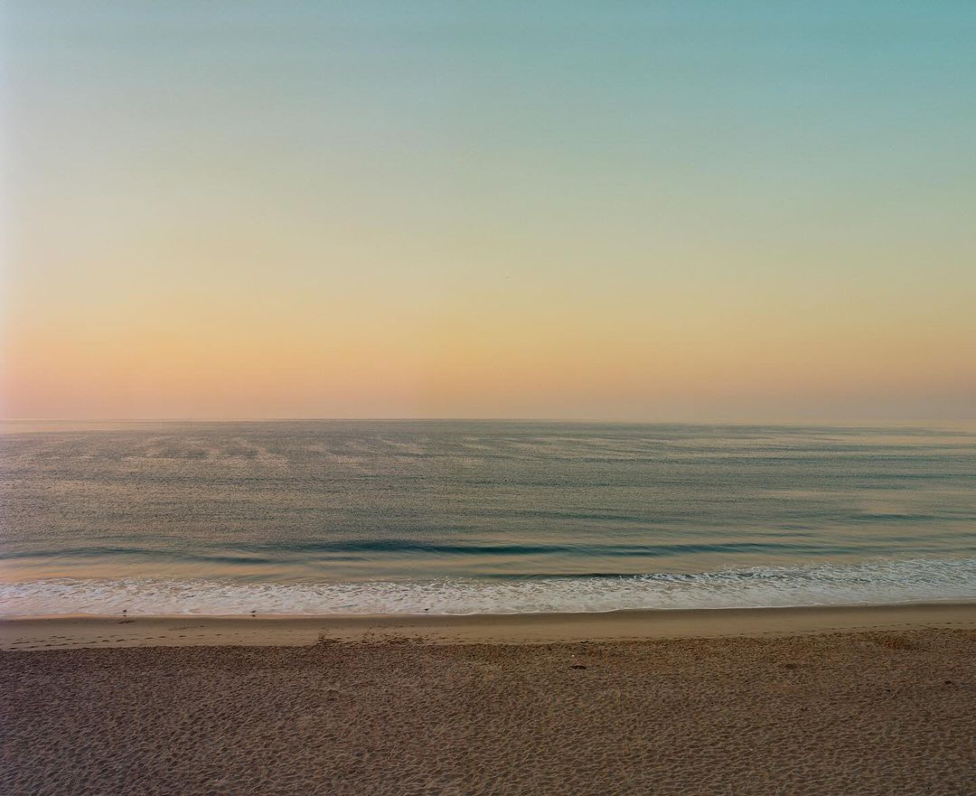 Serene coastal landscape featuring a sandy beach, calm ocean waves, and a gradient sunset sky captured by Rainer Hosch.