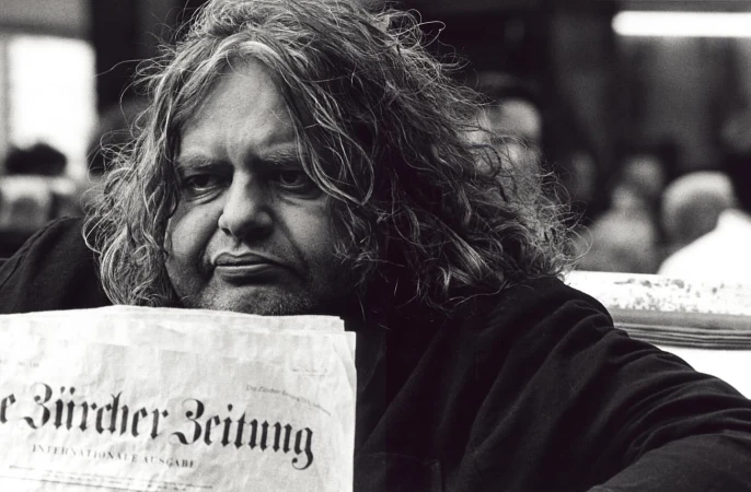 Monochrome portrait of a middle-aged man with long curly hair holding up a newspaper with bold German lettering.