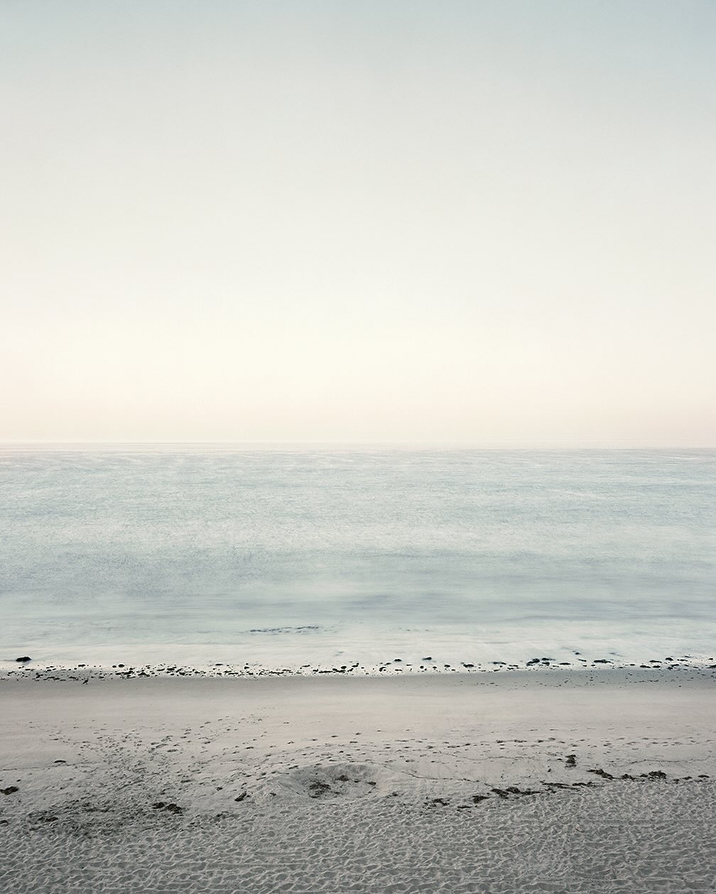 Calm ocean waves gently meet a sandy beach under a serene, overcast sky. Tranquil seascape with muted, natural colors.