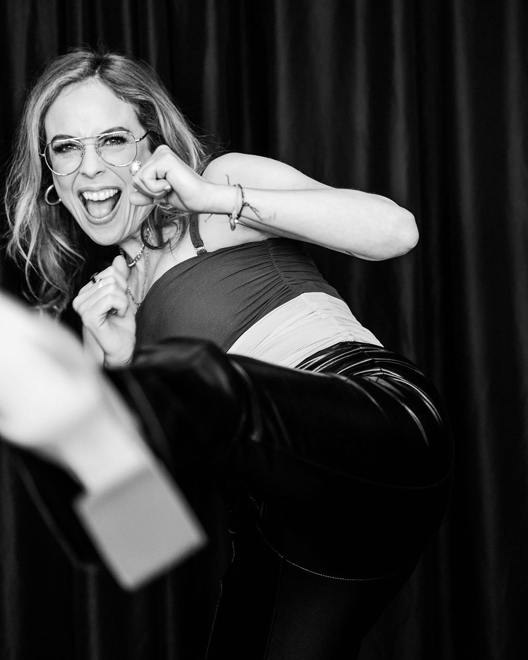 Black and white portrait of an adult woman with glasses in a karate kick pose wearing a strappy top and black pants in front of a curtain.