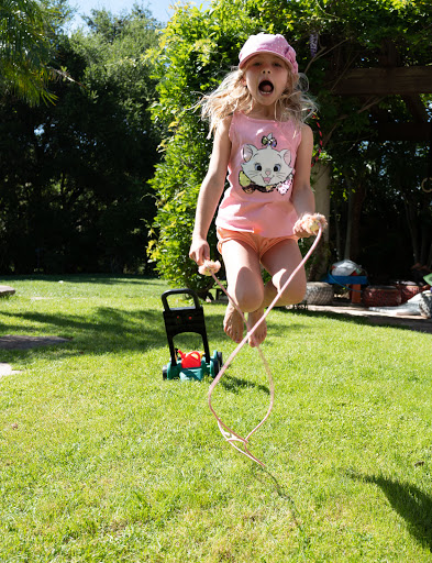 Young girl with a pink hat is jumping rope barefoot in the sun on a green lawn outdoors, with trees and a toy lawn mower in the background.
