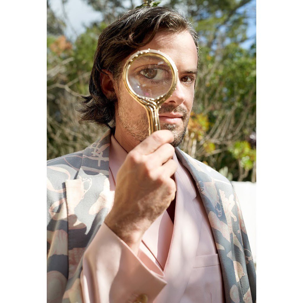 Adult male in floral blazer holds a magnifying glass to his eye outdoors in natural daylight with background foliage and neutral colors.