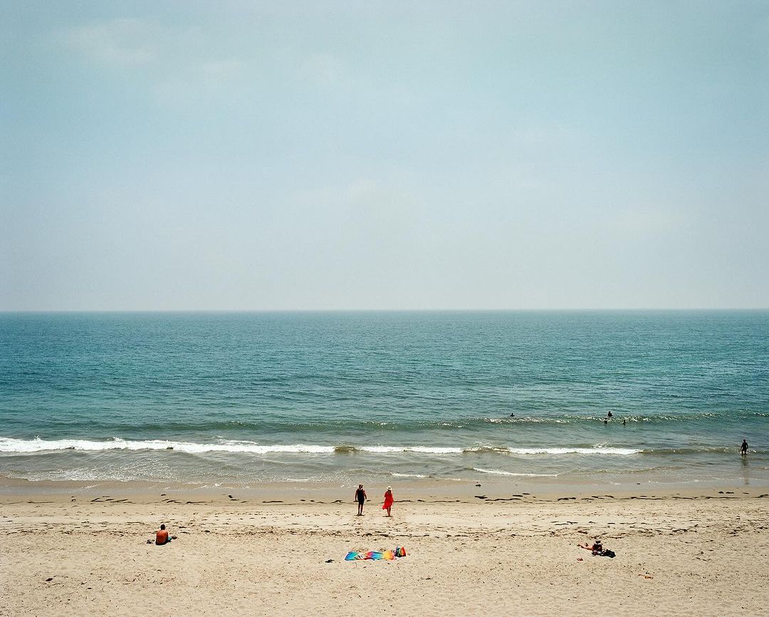 Ocean view on a hazy day shows two people walking on the beach with other people visible in the water and on the sand.