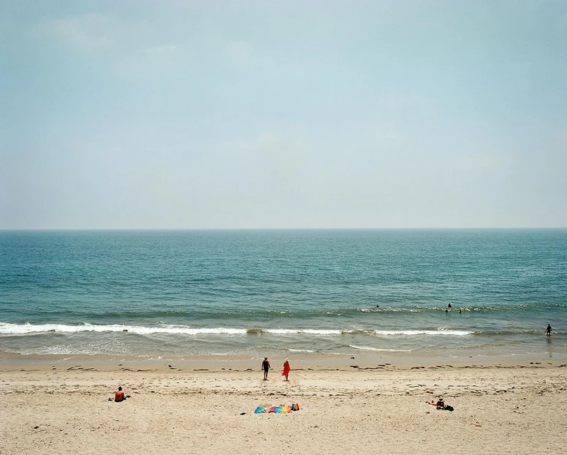 Ocean view on a hazy day shows two people walking on the beach with other people visible in the water and on the sand.