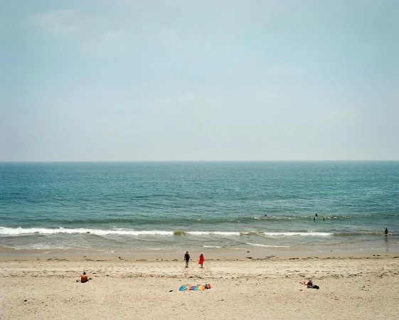 Ocean view on a hazy day shows two people walking on the beach with other people visible in the water and on the sand.