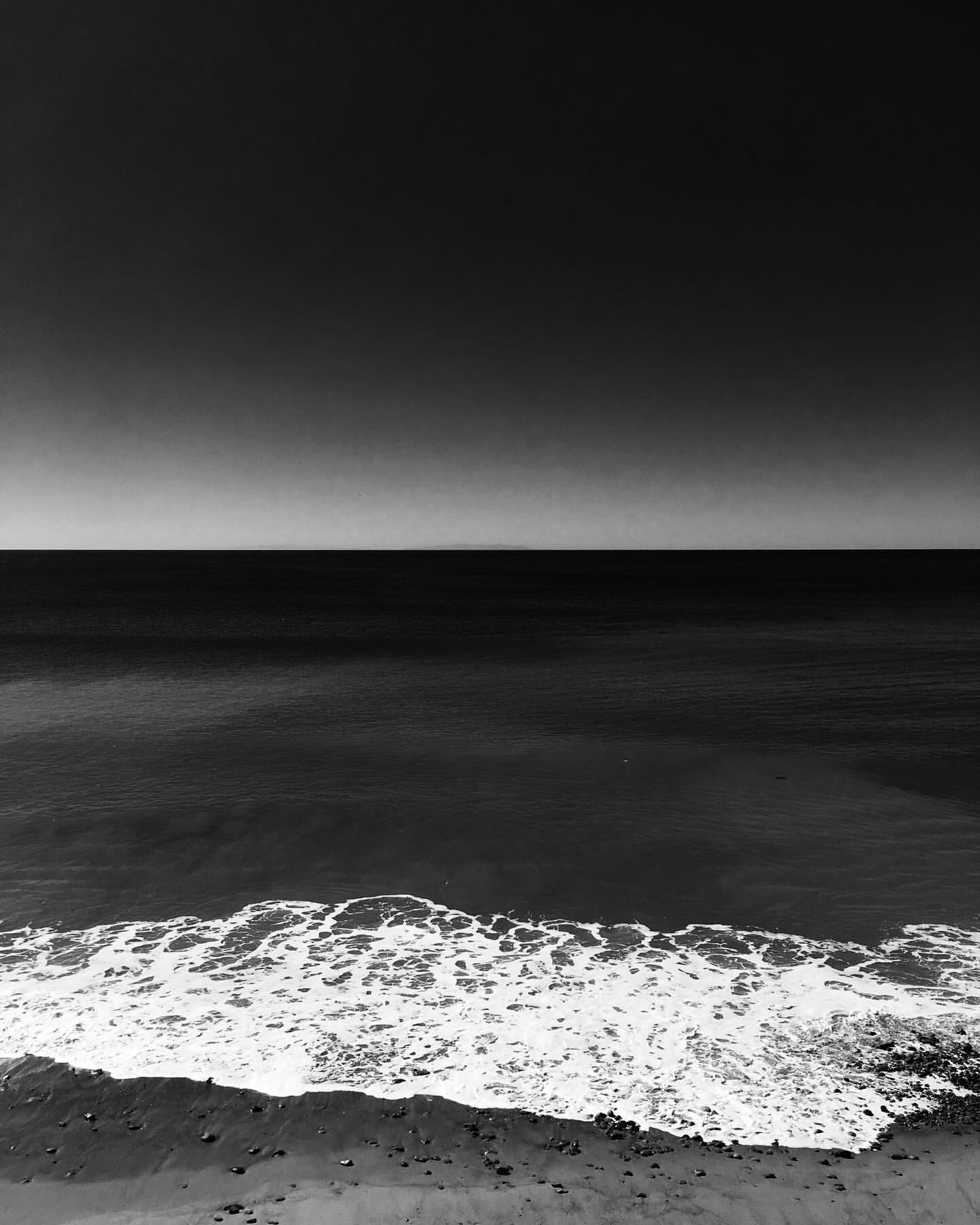 Black and white coastal landscape showing ocean waves breaking on a sandy shore under a wide horizon.