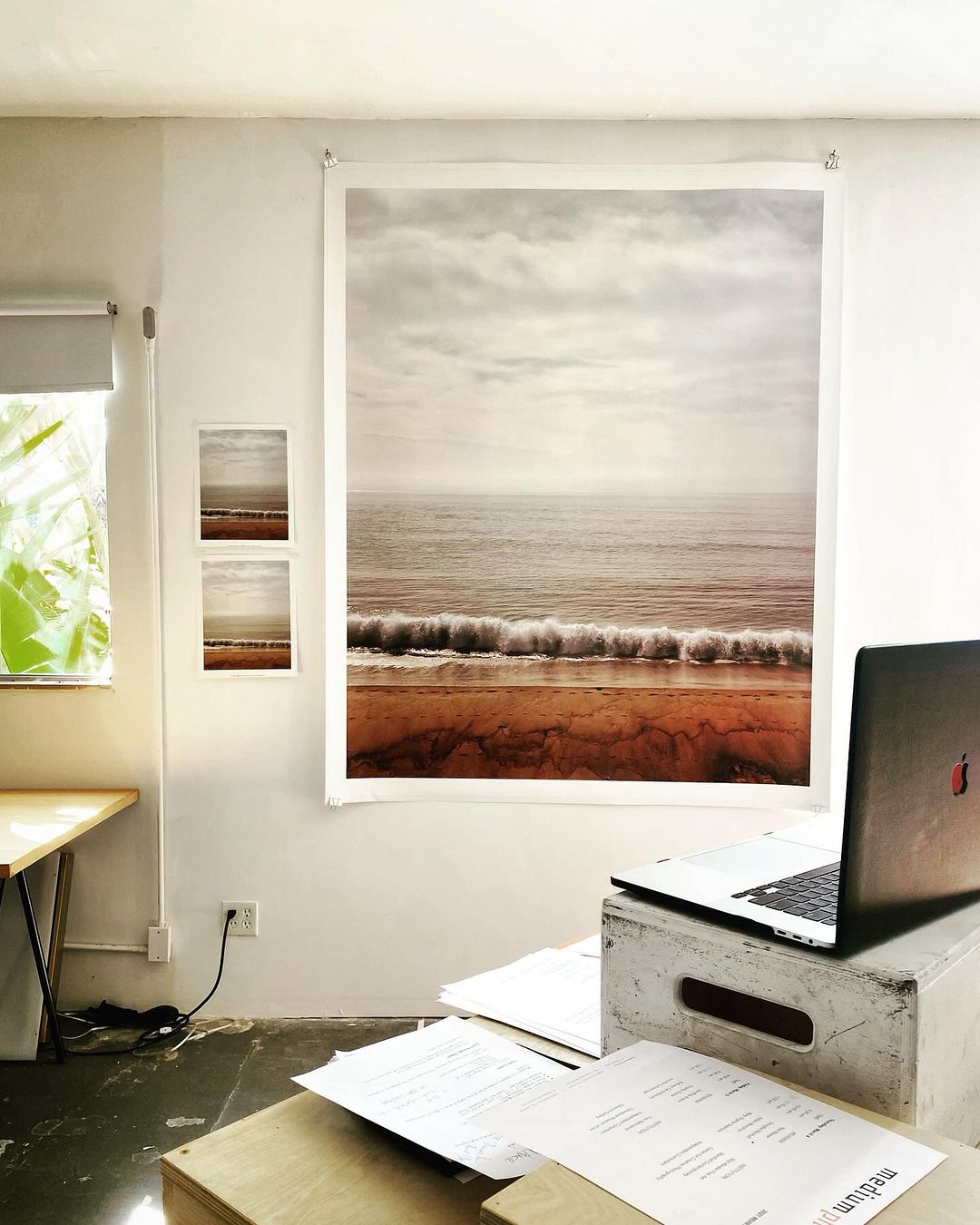 Home office interior featuring a scenic beach landscape photo hanging on the wall, a laptop on a box, and paperwork on a wooden table.