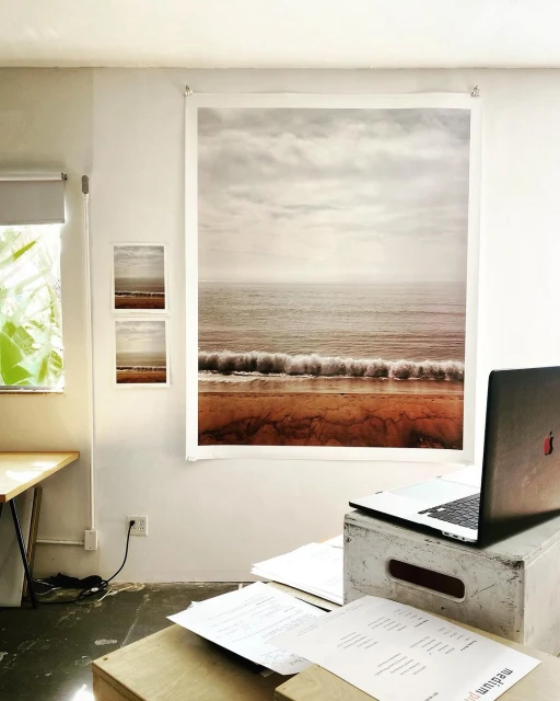 Home office interior featuring a scenic beach landscape photo hanging on the wall, a laptop on a box, and paperwork on a wooden table.