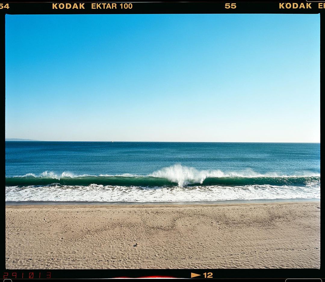 Ocean wave breaks near the beach on a sunny day, capturing the water's movement and the sandy shoreline under a clear blue sky.