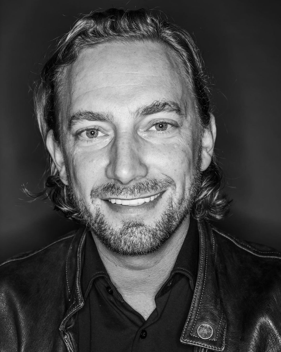 Black and white portrait of a smiling adult male with a beard and medium-length hair, wearing a leather jacket. Head and shoulders shot.