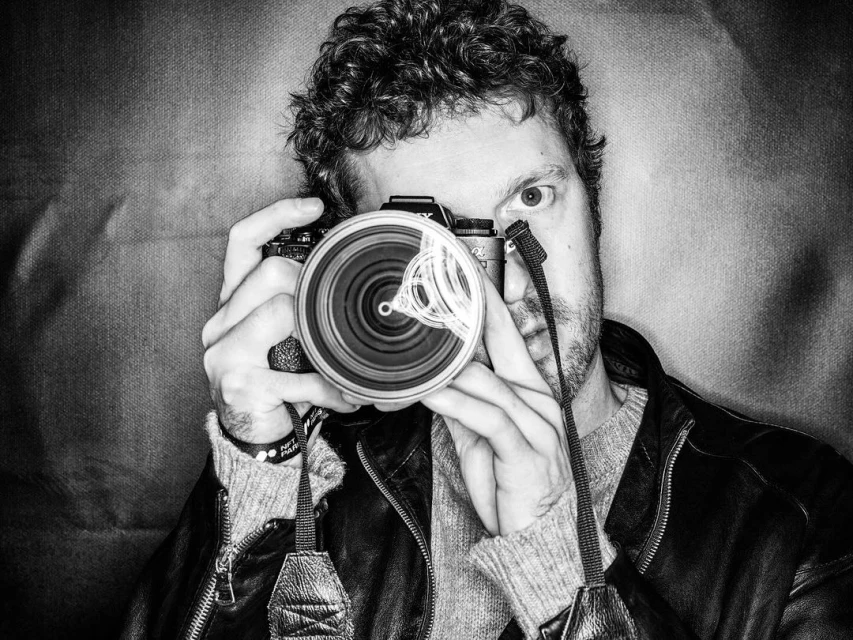 Man holding a camera with a large lens, focusing, in a close-up shot, wearing a leather jacket, against a neutral, textured background.