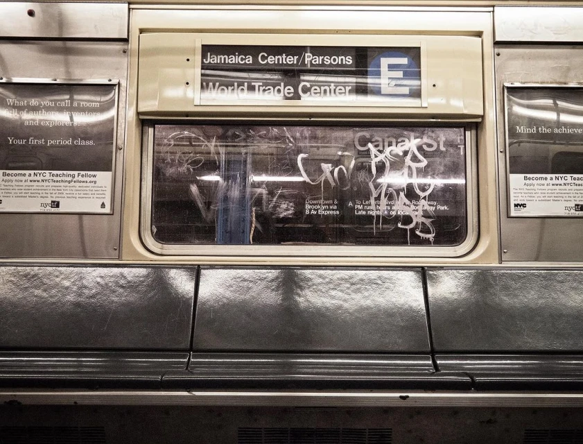 Interior of a subway car featuring signage for Jamaica Center/Parsons, World Trade Center, and the E line, alongside advertisements.