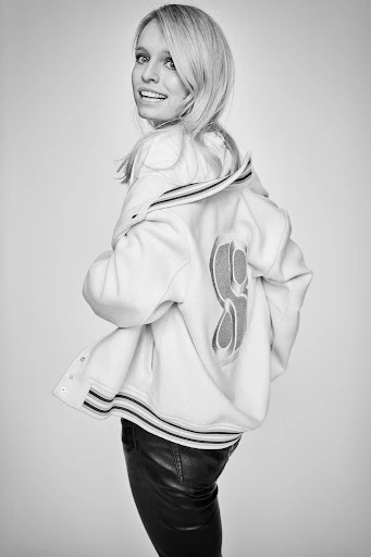 Black and white portrait of a woman with blonde hair, smiling and looking over her shoulder, wearing a letterman jacket and leather pants.