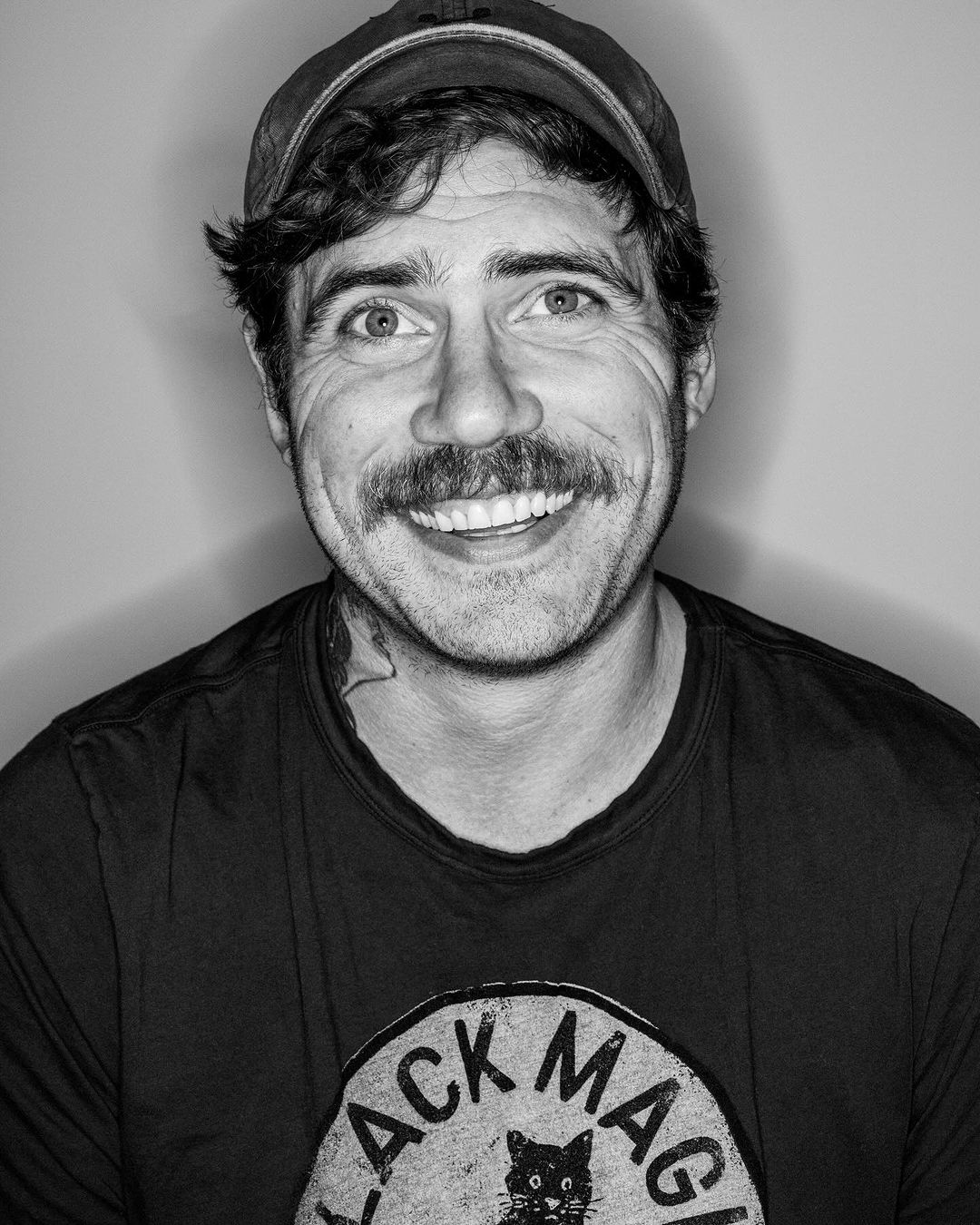 Monochrome portrait of an adult male with a mustache, wearing a baseball cap and t-shirt with a graphic, smiling in a studio setting.