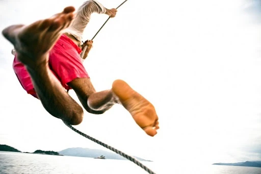 Low-angle shot of a person swinging on a rope over water. Focus on legs, feet, and rope against a bright sky and distant land.