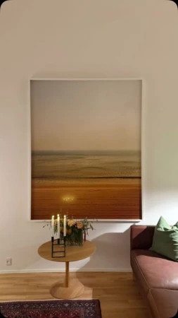 Living room interior featuring a landscape painting, floral arrangement, candles, wooden table, and part of a leather sofa with a green cushion.