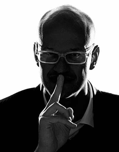 Monochrome portrait of a bald adult male wearing glasses and a suit, holding a finger to his lips in a shushing gesture against white.