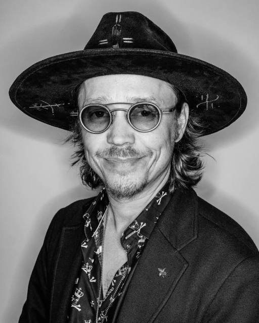 Black and white portrait of an adult man wearing a hat and glasses, with a patterned shirt and jacket, against a plain backdrop.