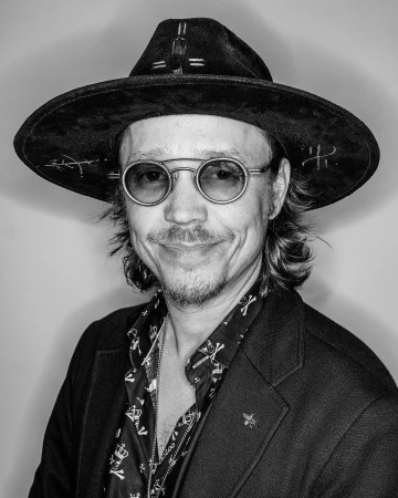 Black and white portrait of an adult man wearing a hat and glasses, with a patterned shirt and jacket, against a plain backdrop.