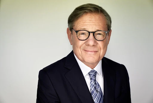 Portrait of an older man with glasses wearing a dark suit and patterned tie against a plain white background.