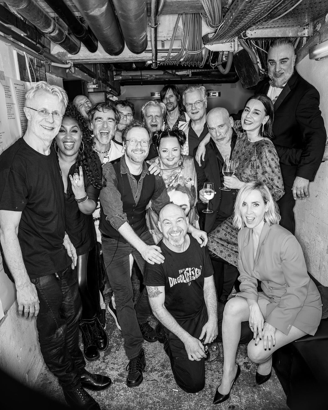 Black and white group portrait of approximately 15 people in an indoor setting, possibly backstage or in a bar. The photograph is well lit.