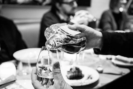 Person pouring a clear liquid from a glass bottle into a stemmed glass at a table with other glassware and people in background.