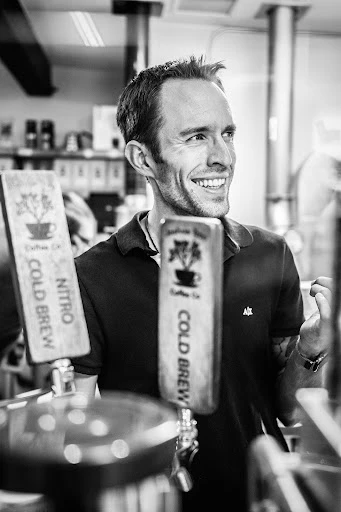 Monochrome portrait of a smiling man in a polo shirt standing near two cold brew taps inside a coffee shop or bar.