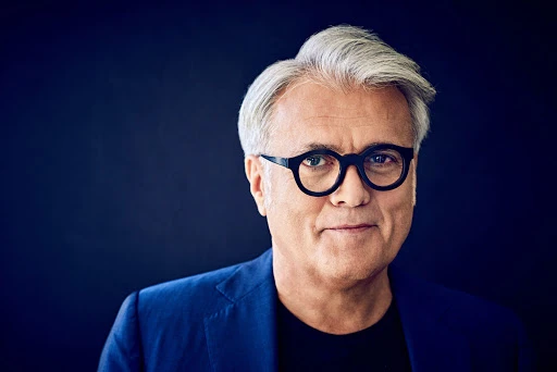 Portrait of a mature adult male with grey hair and glasses, wearing a blue blazer against a dark blue background in a studio.