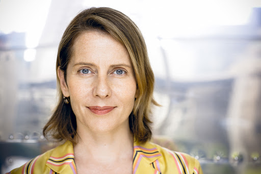 Close-up portrait of a smiling adult woman with light brown hair wearing a yellow striped blazer against a blurry silver background.
