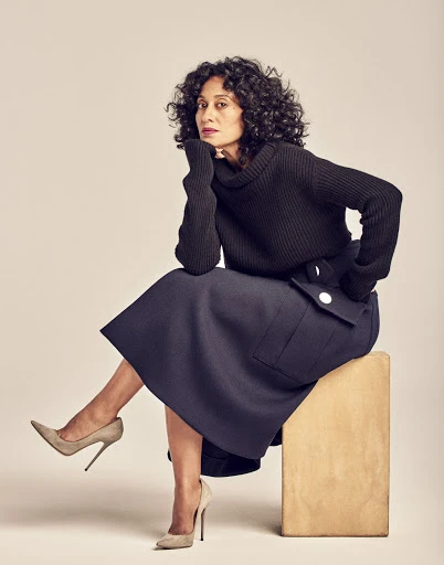 A stylish adult woman with curly hair sits on a wooden cube wearing a black turtleneck sweater and a navy blue skirt against a beige backdrop.