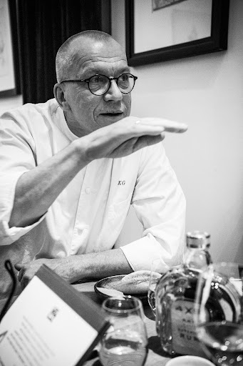 Monochrome medium shot of a mature adult man with glasses, wearing a white shirt and gesturing with his hand, indoors with bottles and a menu.