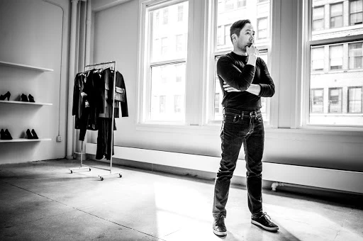 Asian man in casual clothes standing in an urban studio with natural light, clothes rack, and simple white shelving with shoes displayed.