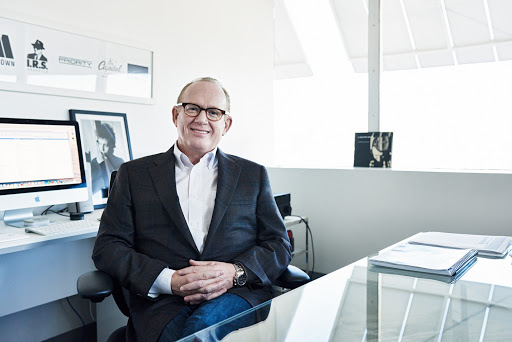 Businessman in glasses wearing a suit seated at a modern desk with a computer, keyboard and framed posters in the background.