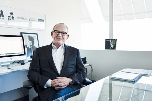 Businessman in glasses wearing a suit seated at a modern desk with a computer, keyboard and framed posters in the background.