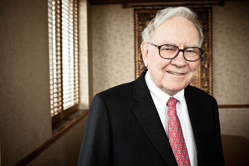 Portrait of an elderly man in a suit and glasses with a red patterned tie, posing indoors with neutral wall and window.