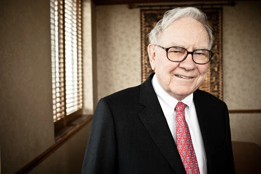 Portrait of an elderly man in a suit and glasses with a red patterned tie, posing indoors with neutral wall and window.