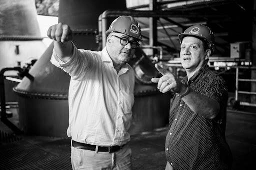 Two men in hard hats point while standing in an industrial setting with pipes and equipment, captured in a monochrome photograph.
